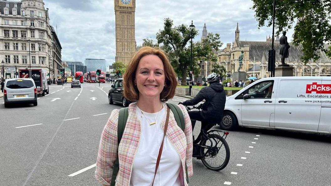 Lola McEvoy standing near Big Ben and Parliament in London. She has brown hair and is wearing a pink checked jacket and white top. There are a lot of red busses in the background while, close to the MP, is traffic including vans and a cyclist on a black bike.