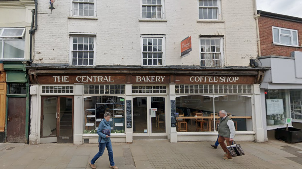 Two people walk in front of a bakery, a white-brick building with a brown wooden sign saying 'The Central Bakery Coffee Shop'