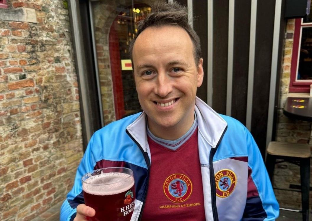 Stewart Garratt, wearing an old fashioned claret Villa shirt and a claret and blue tracksuit top while holding a pint of beer.  He is seated outside by a brick wall.