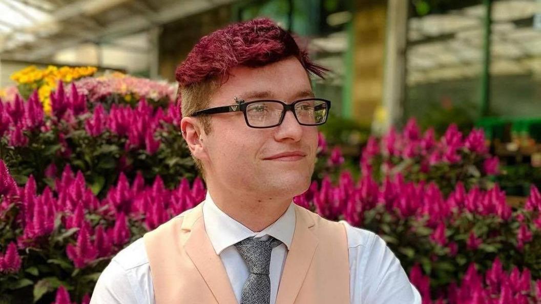 Tommy Parker wearing glasses and a salmon-coloured waistcoat, white collared shirt and grey tie. He is sitting against a background of blurred pink flowers