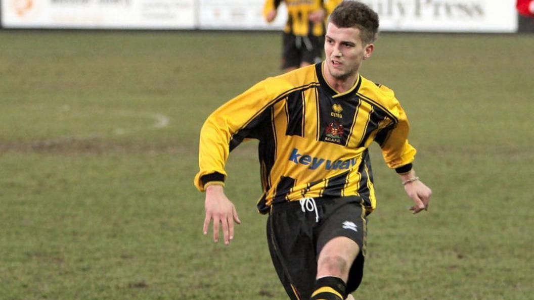 Richard wearing a yellow and black top and black shorts playing in a football match.