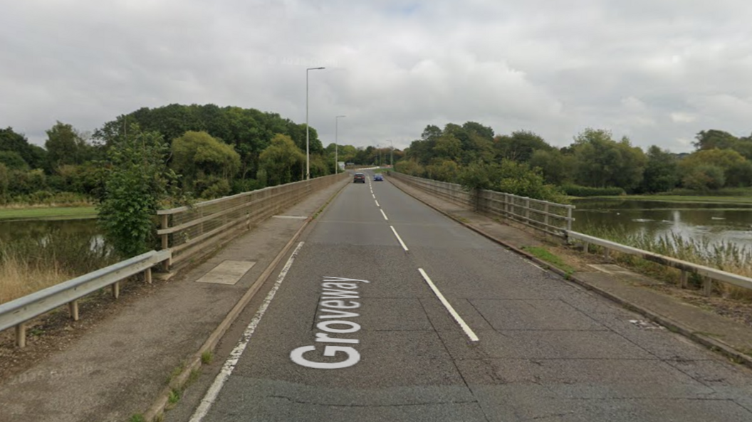 A street view of a road bridge over a river with a footpath on each side with railings. There are two cars travelling on each side of the road in the background.