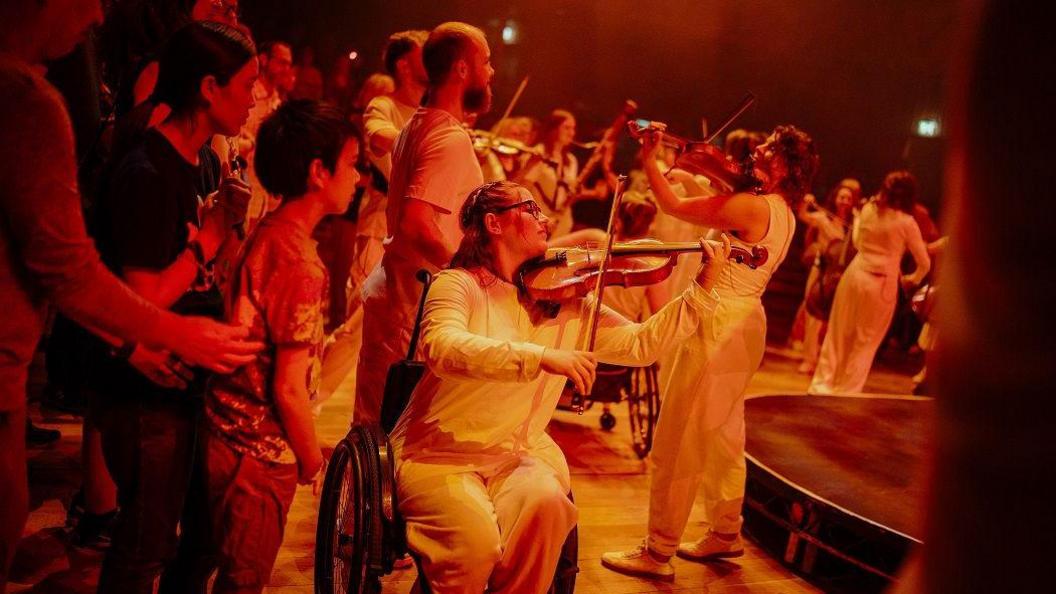 Group of musicians in the background playing, the shot is focused on a woman sitting in a wheelchair playing a violin, dressed in white