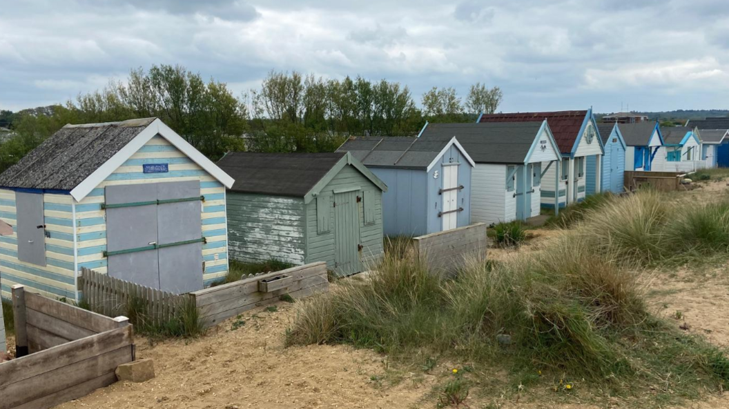 Nine beach huts are lined up on the beach in Heacham. They are either painted green or blue, and all have doors at the front. Sand is in front of them and behind is a row of trees.