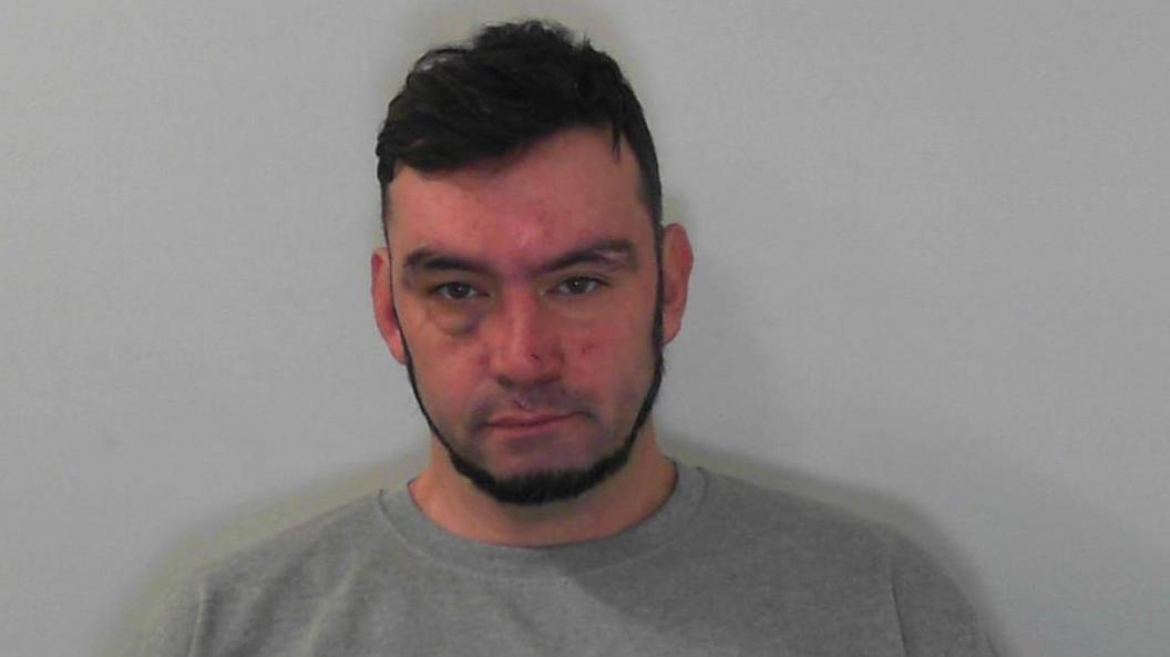 A man with dark hair and a short cropped beard stares at the camera in a police mug shot.