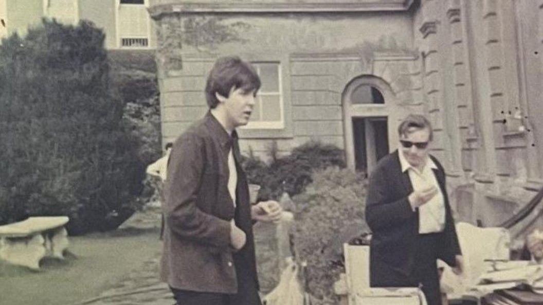 Sir Paul McCartney is in the middle of the photo outside a building, with another man on the right.