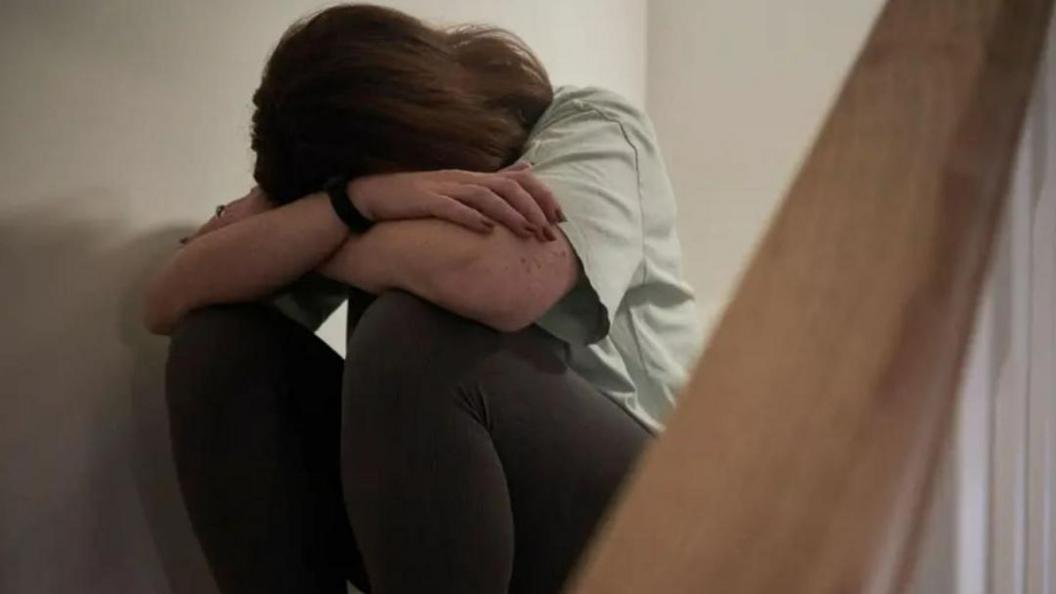A woman sits on a staircase with her hand in her hands, she is wearing a white t-shirt and black trousers