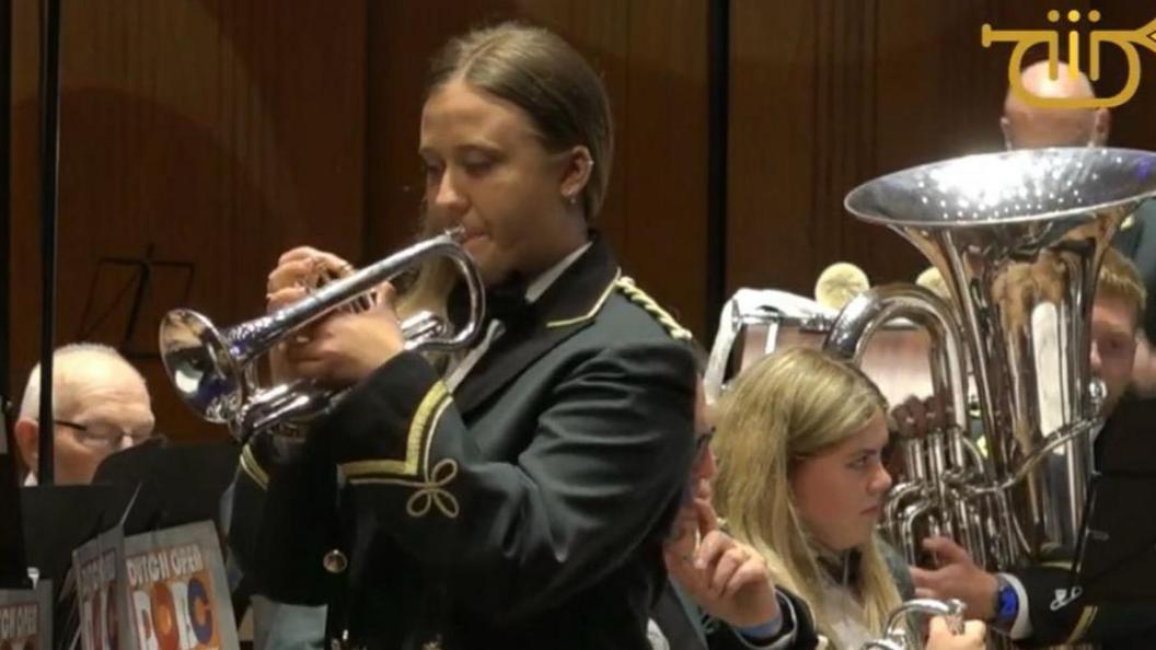 A young girl is pictured from the waist up. She is wearing a white shirt and green blazer with gold threaded embellishments. She has long brunette hair and is holding a silver cornet to her mouth. She is standing in front of a brass band - a large tuba and a few other band members can be seen behind her.