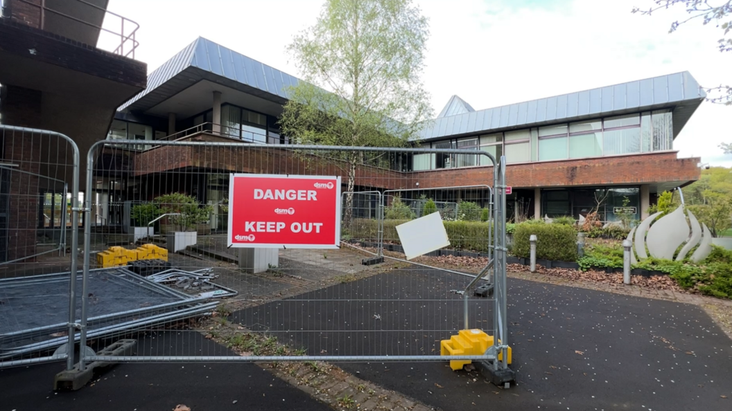 A side entrance to County Hall is blocked by metal fencing and a red sign reading 'Danger Keep Out'. Behind it are slightly overgrown flowerbeds and bushes in front of red-brick walls.