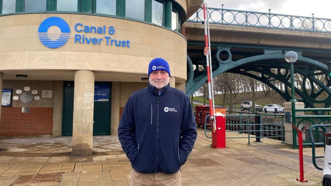 Head and shoulder view of a man with a grey beard wearing a blue beanie and a blue fleece. To his right is the entrance to building in sand coloured stone with a blue sign stating Canal and River Trust and a round blue logo. Above him to his left is a bridge with green metal supports and an ornate green metal railing. Beneath it some cars are parked on a tree-lined road.