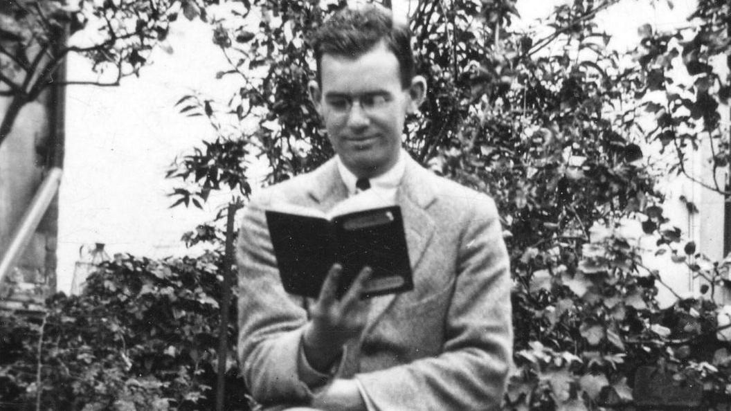 In black and white, a black-haired Gareth Jones wearing a suit and sitting on a stool in a garden near a stone wall, looking at his passport. Behind him are the branches of a plant.