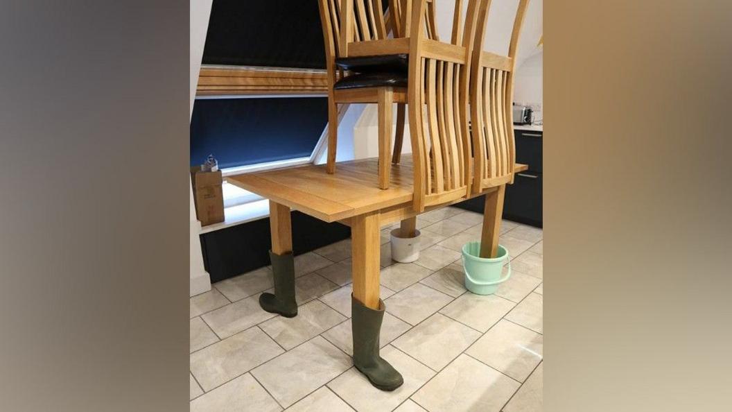 A kitchen table stands on a tiled floor with two legs in welly boots and two legs in buckets. The chairs have been placed on top of the table to keep them out of expected flooding.