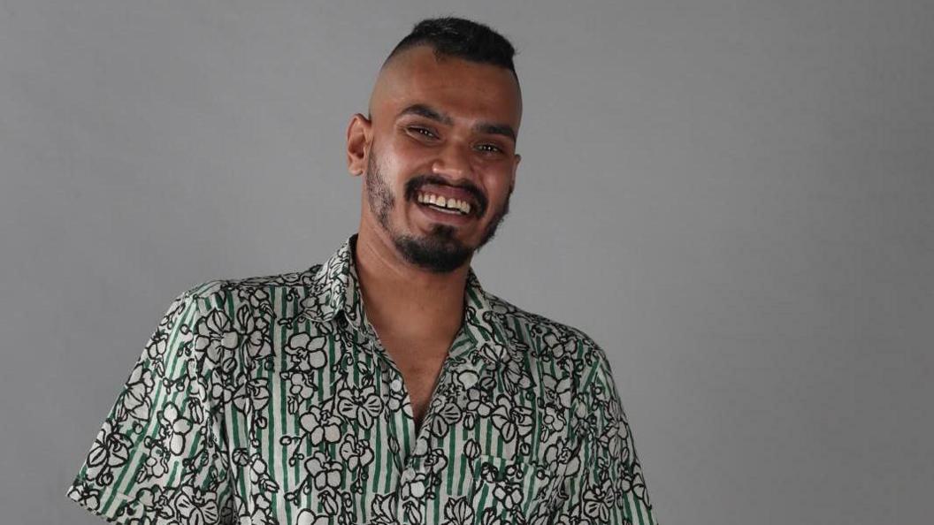 Mohaimon with a big smile looking at the camera, wearing a cotton printed shirt which is both striped and has flower images on it.