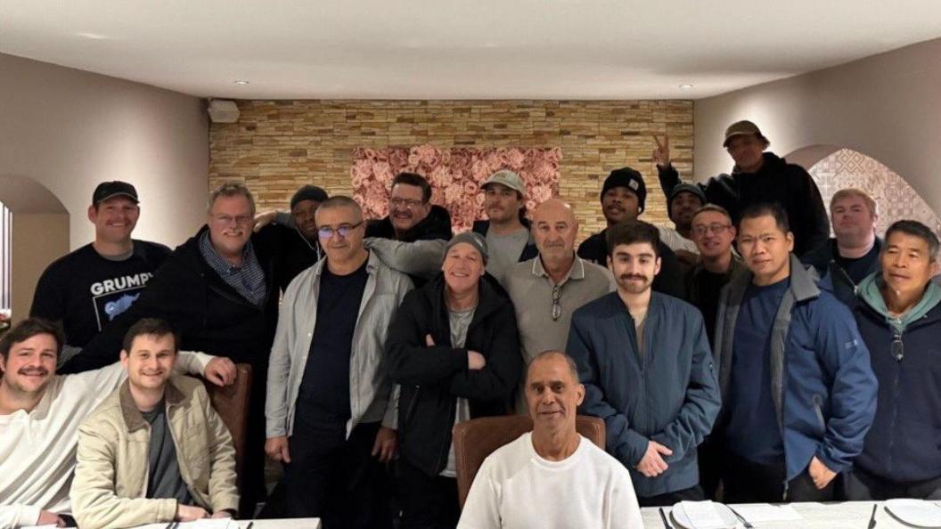 Nineteen men, of various ages, look jubilant as they pose for a photograph. One person, who is at the back and on the left-hand side, is making a 'V' for victory or peace sign.