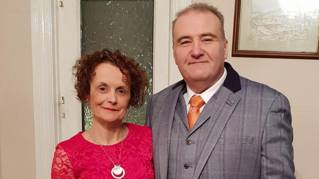 Lisa Castleton and her husband Lee. Lisa has brown, curly hair and is wearing a magenta-coloured dress and a silver necklace, while her husband is wearing a silvery-grey, pin-striped suit and orange tie.