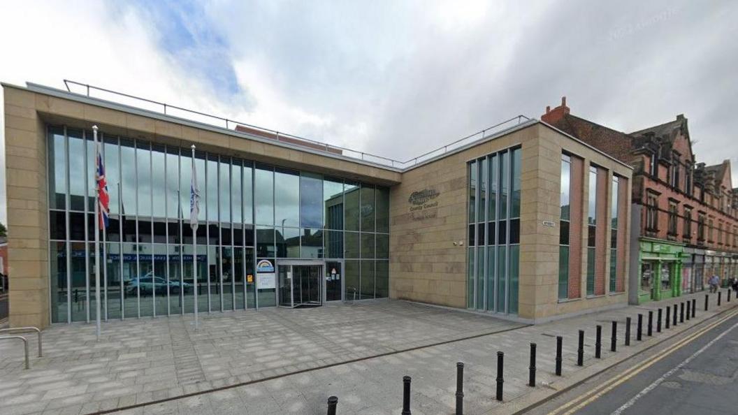 Cumberland Council's headquarters in Carlisle: a two-storey building with high windows. There is a small square and two flags at the front, including the Union Jack. 