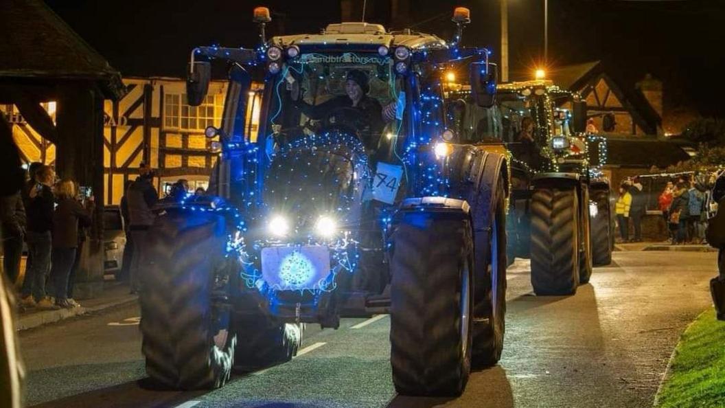 A tractor covered in brightly coloured fairy lights in the middle of the road. Two other tractors can be seen behind it.