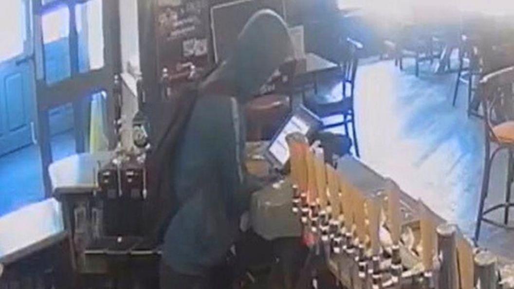 A CCTV image of a man wearing a hooded jumper facing away from the camera. He is behind the bar of a pub and trying to access the cash register. 