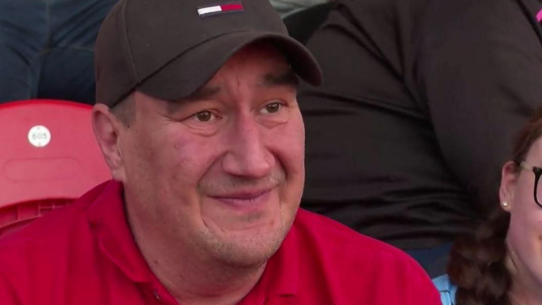 Ady Good wearing a black cap and a red polo shirt is sat in a crowd at a stadium