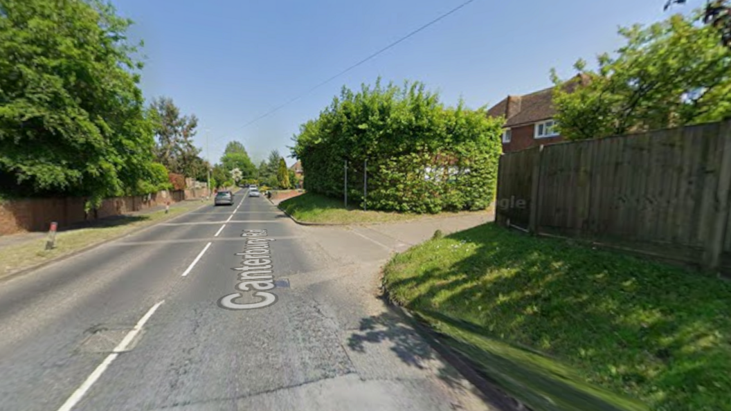 A tarmac road with few cars on it. There is a turning off the road into an area with a brick house. There is a hedge on one side of the turn-off and a high, wooden fence on the other.