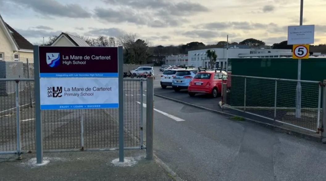 The entrance to La Mare de Carteret school, which has a sign and a gate leading to a car park with four cars visible. 