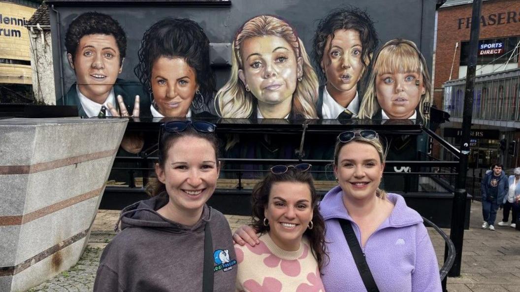 Three friends - Annie O'Connor; Rebecca Mueller and Niamh Dalton - standing in front of the Derry Girls mural. 