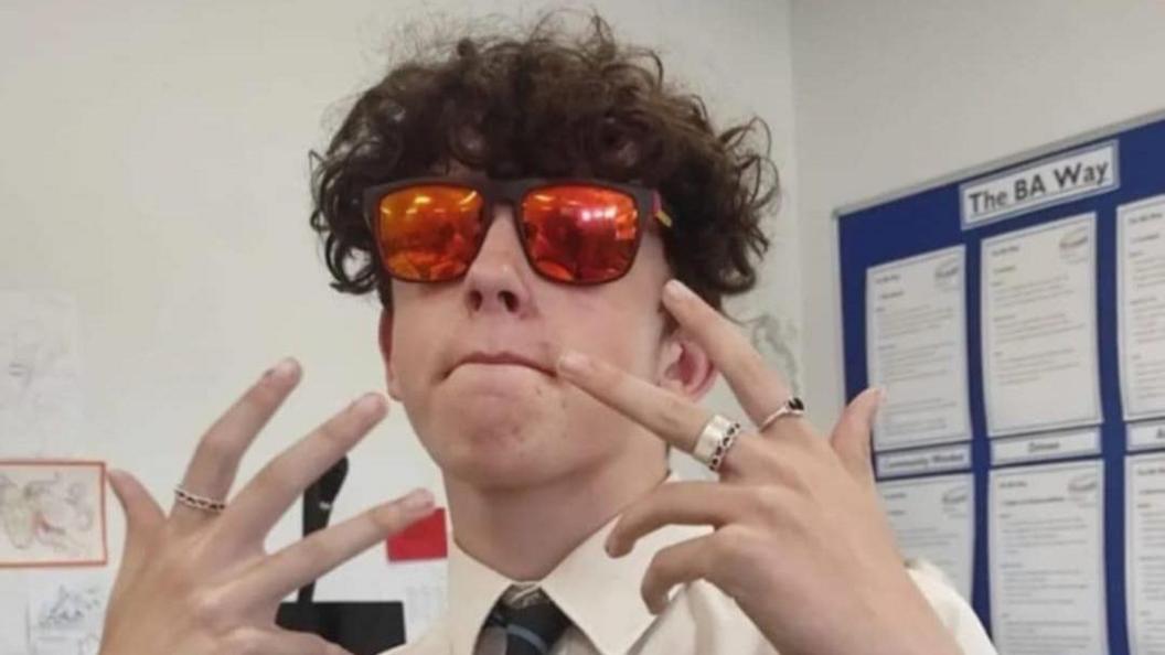 Thomas Taylor is posing for the camera, wearing a pair of red or orange-tinted sunglasses. He has rings on his fingers and is wearing a white shirt with what looks like a striped school tie. Thomas has dark, wavy hair