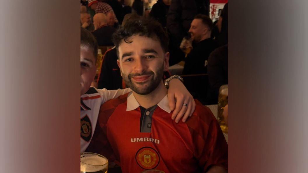 A man with black, curly hair, and a beard, looks directly at the camera. He is wearing a red football shirt bearing the word 'Umbro', and a football emblem underneath. Another man has his arm around his shoulder. Crowds can be seen behind them.