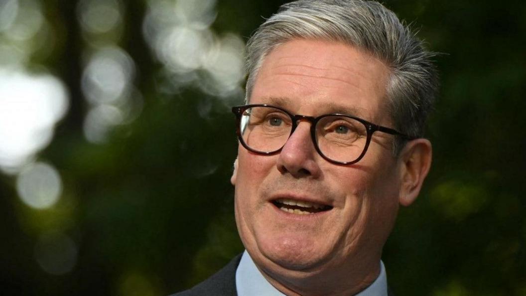 Keir Starmer stands facing the viewer looking to his right. He is wearing glasses and wearing a dark suit with light blue shirt and patterned silvery tie.