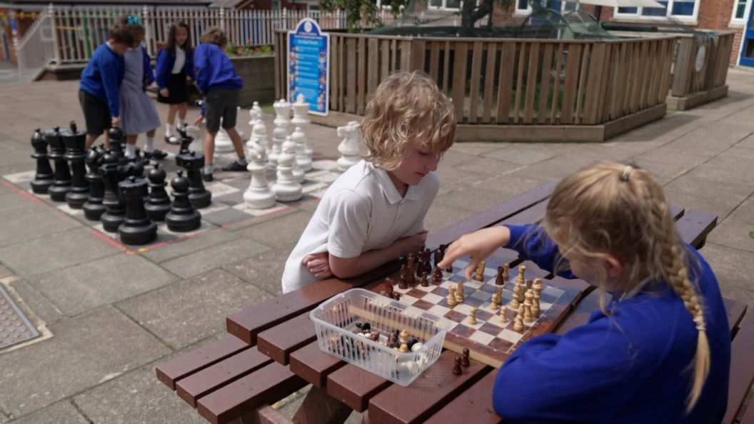 Two pupils are sat on a brown bench in a school playground playing chess on a wooden board. One girl has light blonde hair and is playing white, she is making her move. Opposite her is a young boy with long blonde hair playing with the brown pieces. Behind them is a set of large chess pieces on the school playground, four children are moving the pieces. 