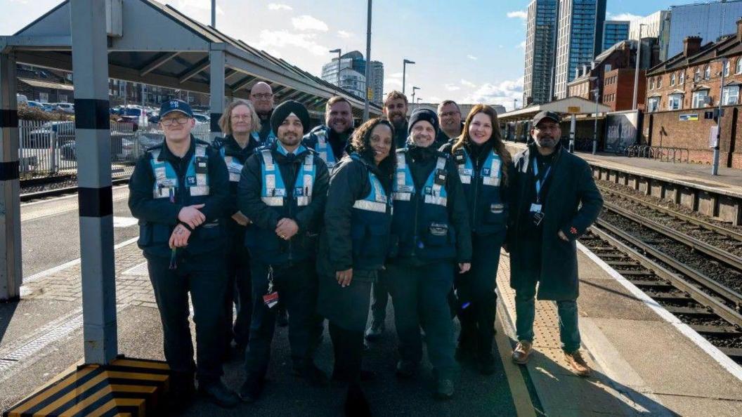 A group of 11 South Western Railway revenue officers, all wearing blue uniforms and blue hi vis vests, pose together on the platform of Woking station.