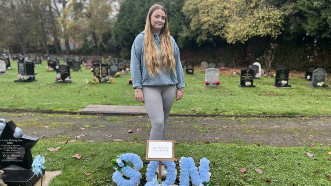 A blonde woman is wearing a grey hoody jumper and grey leggings. She is standing behind a blue wreath that says son, in a blue floral arrangement 