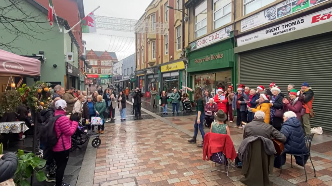 People stood watching a choir, who are wearing Christmas hats, on Mill Street