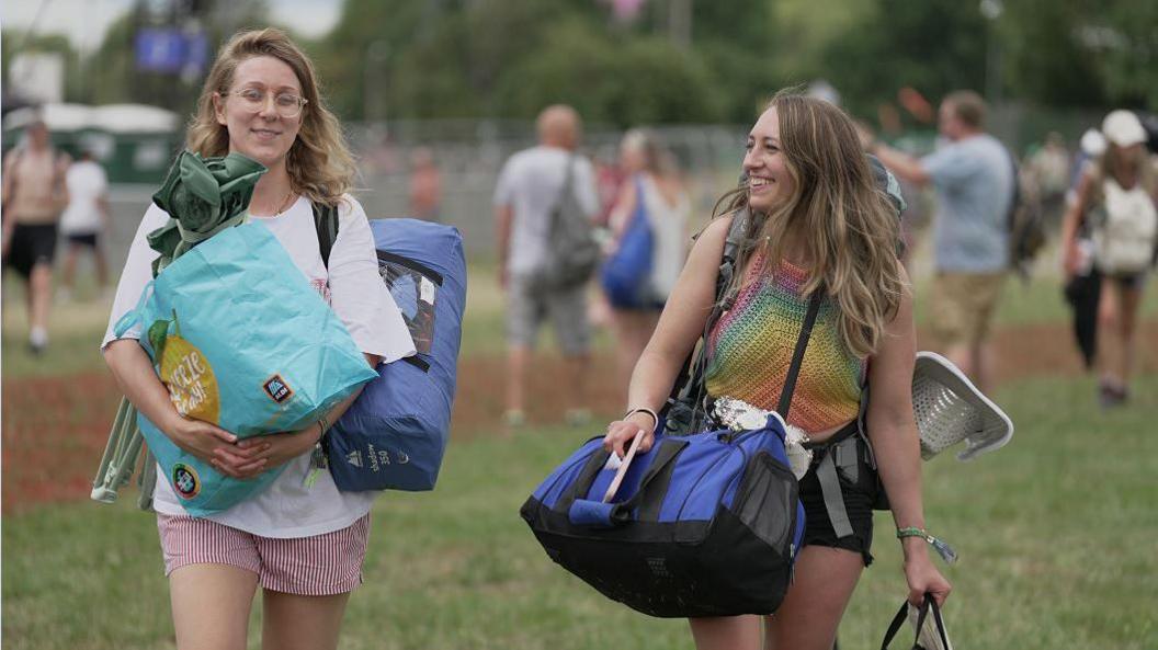 Girl smiles at friend whilst walking through a field carrying their luggage
