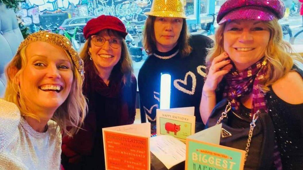 Four women with party hats on sit around a table with birthday cards on.