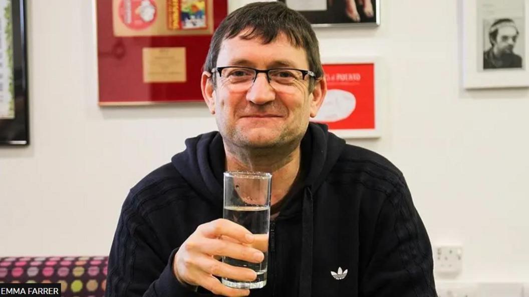 Paul Heaton sitting holding a drink of clear liquid. He is wearing glasses and smiling. There are pictures on the wall behind him