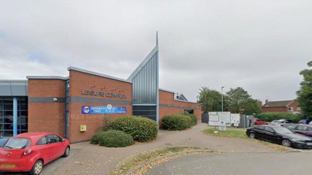 A building is on the left and in the middle of the photo. The words leisure complex are on the outside of the building and a mainly blue sign is below those words. A red car is on the left and other vehicles are on the far right of the image.