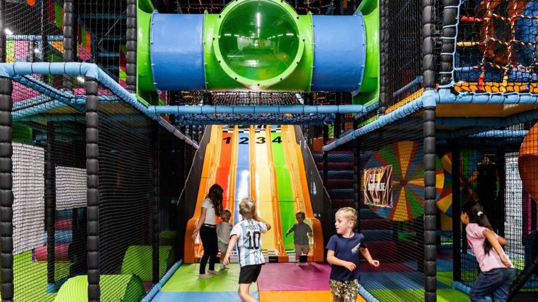 A picture of children playing in a brightly coloured soft play. There is a slide in the background and a tube for crawling through.