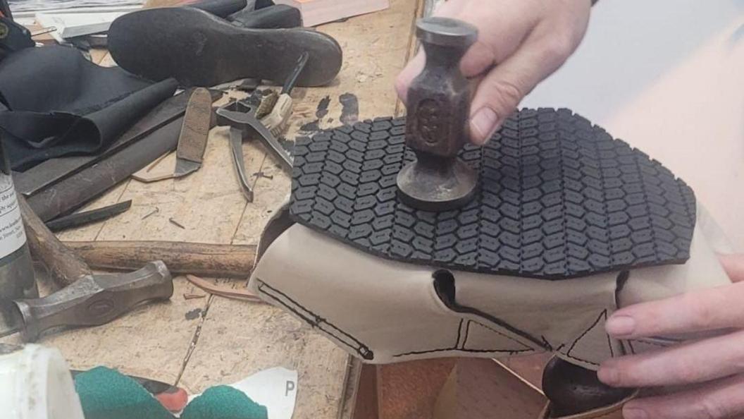 Stuart Moss making the shoes for the camel. He is using a hammer to attach the black rubber soles to the boots, which are white and made of leather and memory foam. 