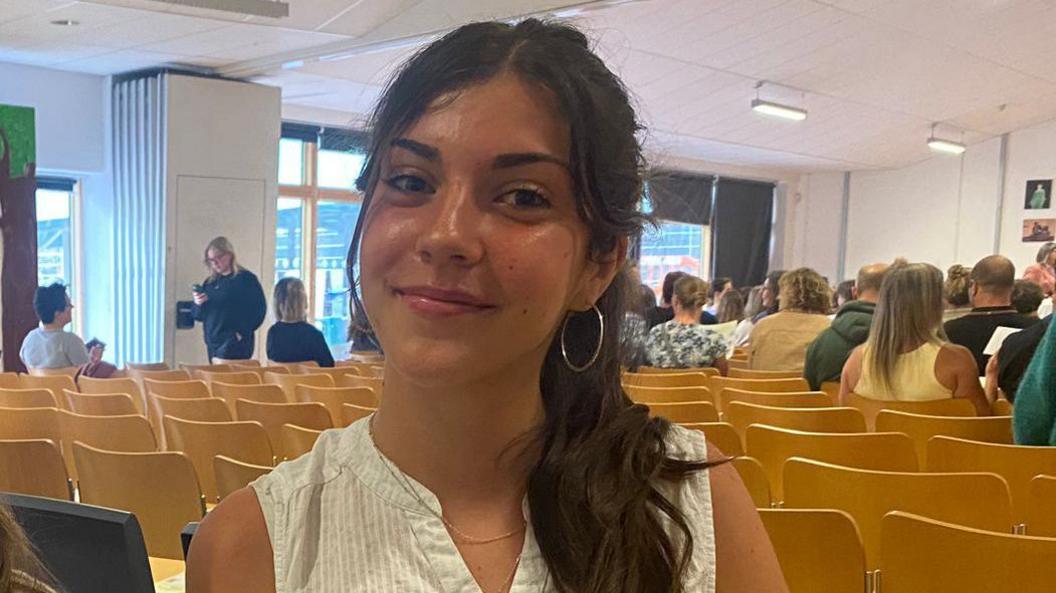 Sofia sits in a school hall. She has long, straight dark-brown hair which is tied up in a ponytail and is wearing a white sleeveless top.