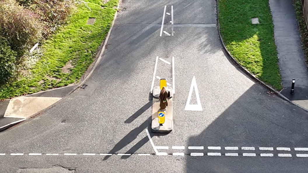 A newly painted t-junction shows a woman crossing the road using a pedestrian refuge - a crossing point before the junction. She stands in the centre of the junction looking out for any traffic. Grass verges can be seen either side of the road.