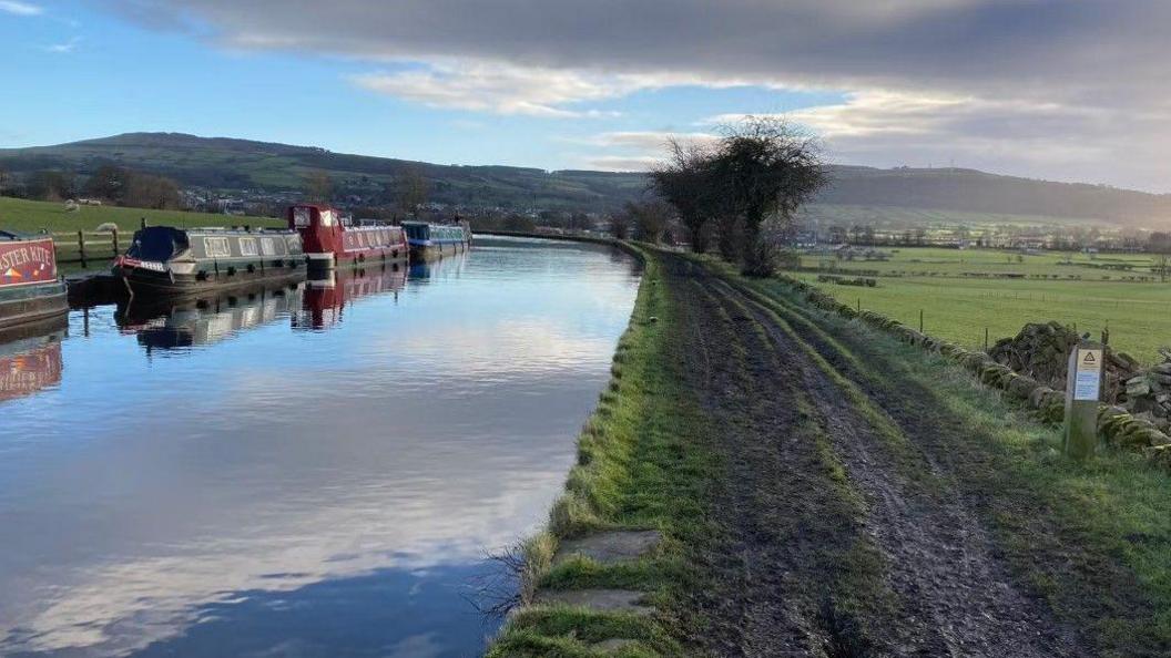 A canal with several barges on the water. Running alongside the water is a muddy pathway surrounded by greenery and hills in the background. The sky is blue with some clouds. 