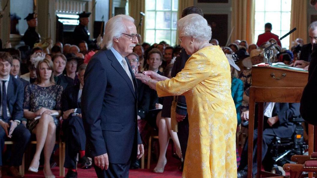 The Queen pins an OBE medal to Charlie Miller. She is wearing a yellow dress and he is in a dark suit and white tie.