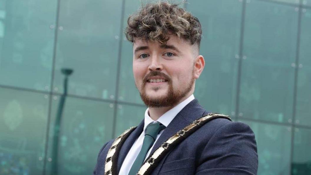 Bréanainn Lyness wearing a suit and tie and ceremonial chain. He is smiling into the camera. Behind him a large glass building.