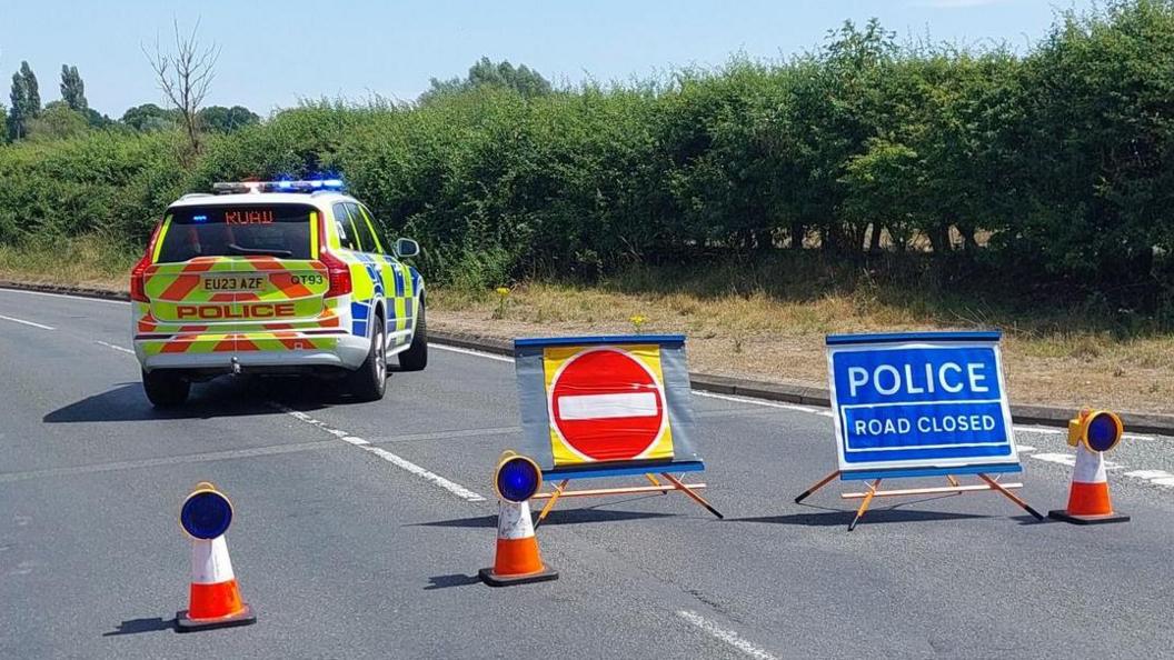 A four-by-four police car blocks the road, which has a thick green bush next to it. There are cones and signs advising that the road has been closed. The picture was taken on a sunny day.