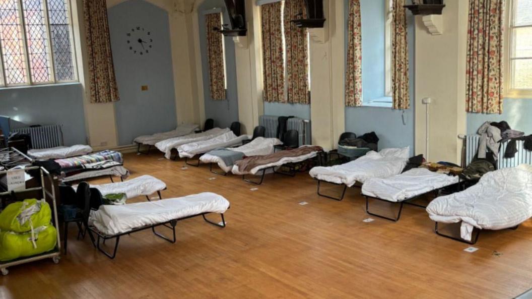 A number of made up camp beds in a room, with pillows and white duvets on them. There is a radiators in the room, with clothes on them, blue walls, patterned curtains, a wooden floor and a clock on the wall. Some personal items are by some beds.