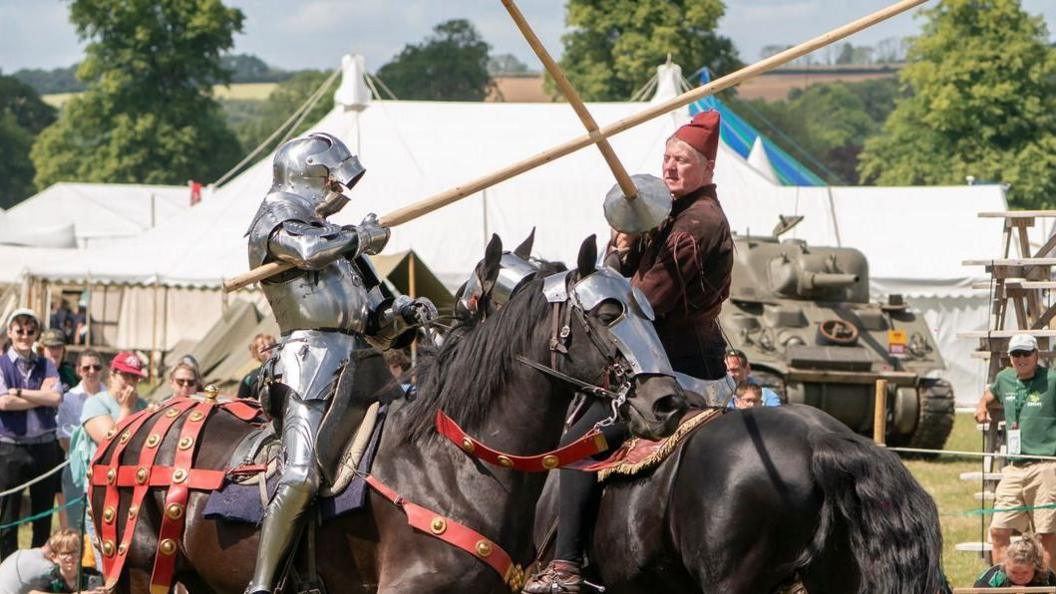 Men on horseback, one in full armour. They are re-enactors, the horses are wearing traditional tack for a demonstration of fighting with lances or jousting. A fence surrounds, which spectators are watching from with the roofs of numerous marquees in the background