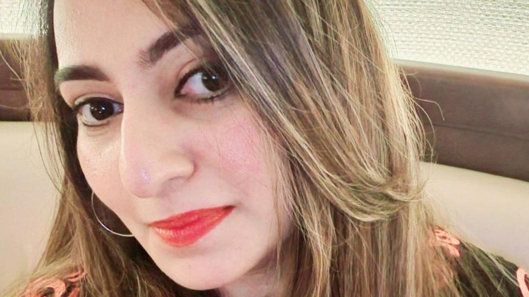 Yaditi Kava has long blonde hair and red lipstick. She is wearing a black and pink dress.