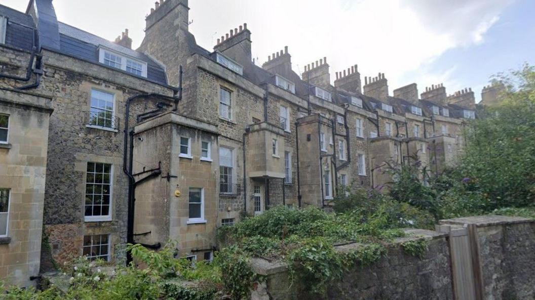 A row of three-storey brick terraced houses in Bath. They are mostly built using the Cotswold stone style of the area and there are plants and bushes running along a wall in front of the properties