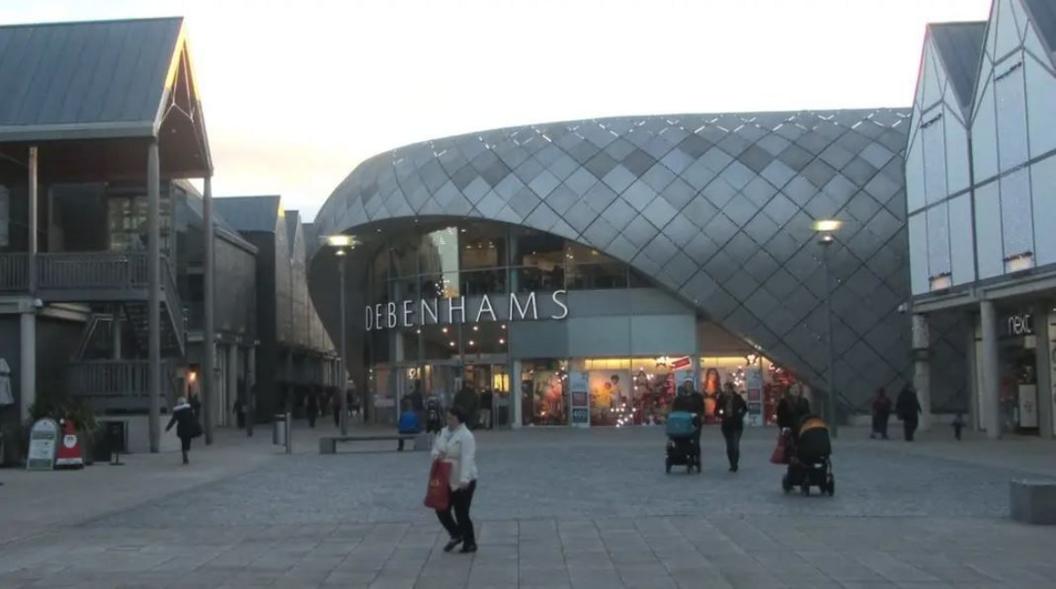 The former Debenhams store in Bury St Edmunds. It has a curved, lozenge-shaped roof clad in grey diamond shapes. Under the roof is the glass-fronted entrance and first floor of the store. Shoppers are walking around on the pedestrianised area in front of the store with other buildings bordering.